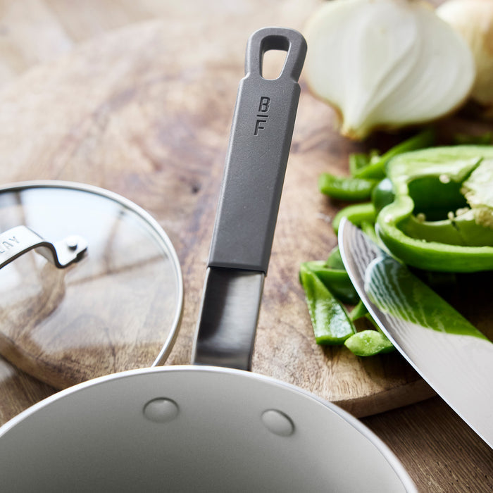 Ceramic nonstick saucepan with lid, assorted vegetables, and knife on a wooden cutting board.