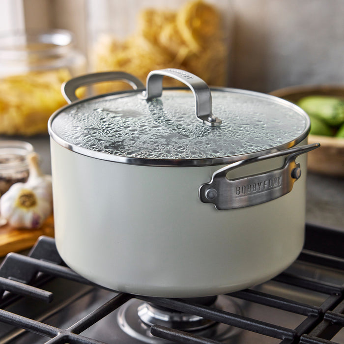 Green ceramic stockpot with lid on stovetop, with dry pasta and garlic cloves beside it.