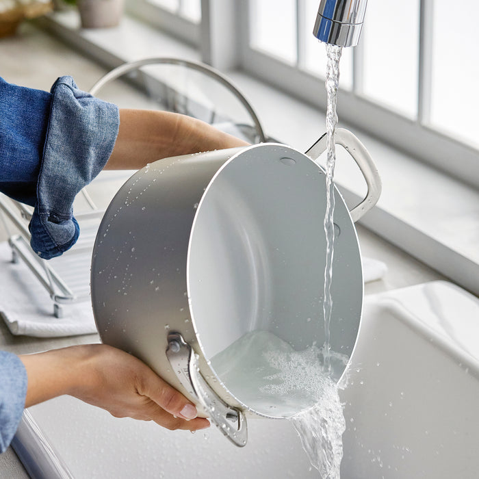 Person rinsing a silver ceramic stockpot under a kitchen faucet near a sunlit window.