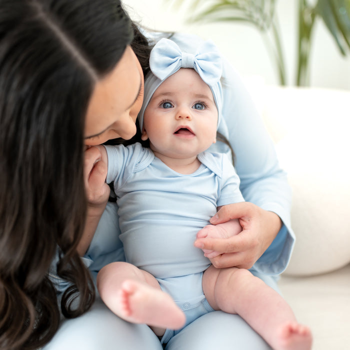 Baby being held by mom, wearing short sleeve bodysuit in a light blue color with matching bow