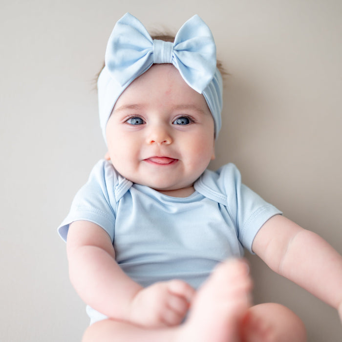 Infant girl laying on a light neutral surface wearing the Bow Headband in Breeze with matching short sleeve bodysuit