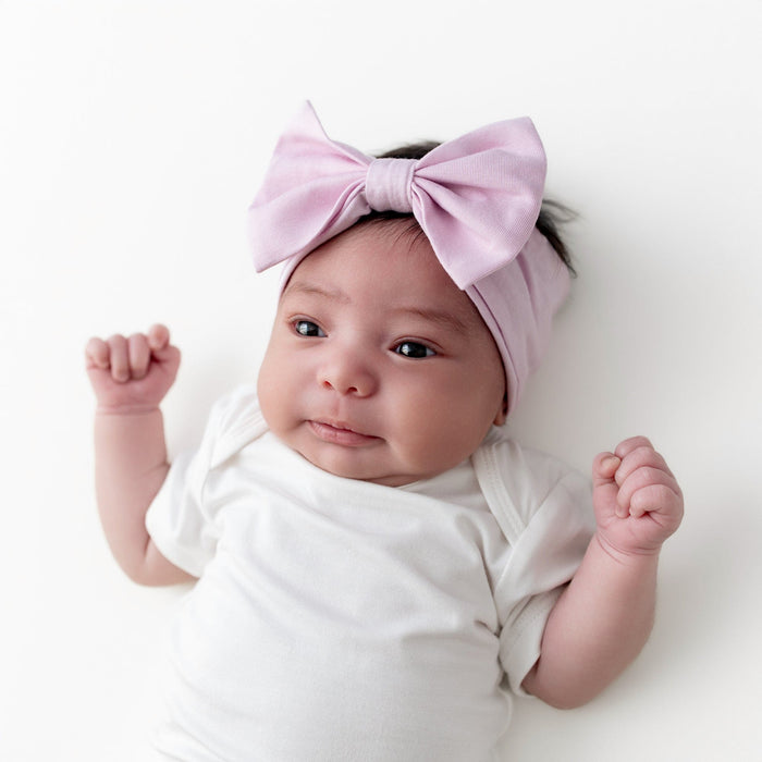 Infant laying on a white surface wearing the Bow Headband in Thistle with a cloud short sleeve bodysuit