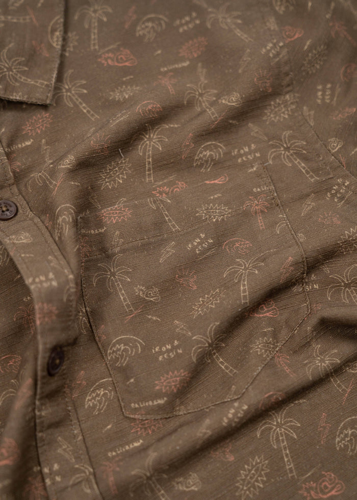 Brown shirt with palm tree and sun patterns, featuring a pocket and buttons. Casual, tropical design.