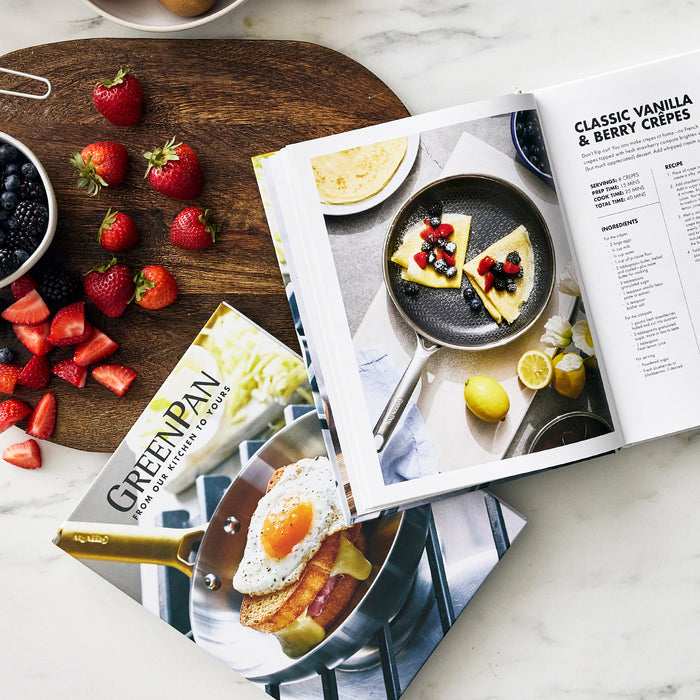 Open to a crepe recipe with a photo of berry-topped crepes in the Cook's Journal Cookbook, Vol. 2, beside a GreenPan catalog and a board holding strawberries, blackberries, blueberries, and eggs on a white countertop.