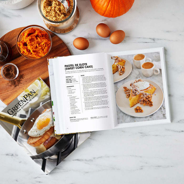 The Cook's Journal Cookbook, Vol. 2 is open to a sweet corn cake recipe on a marble counter, surrounded by eggs, pumpkin puree, and spices—perfect for discovering new dishes and enjoying PFAS-free cooking.