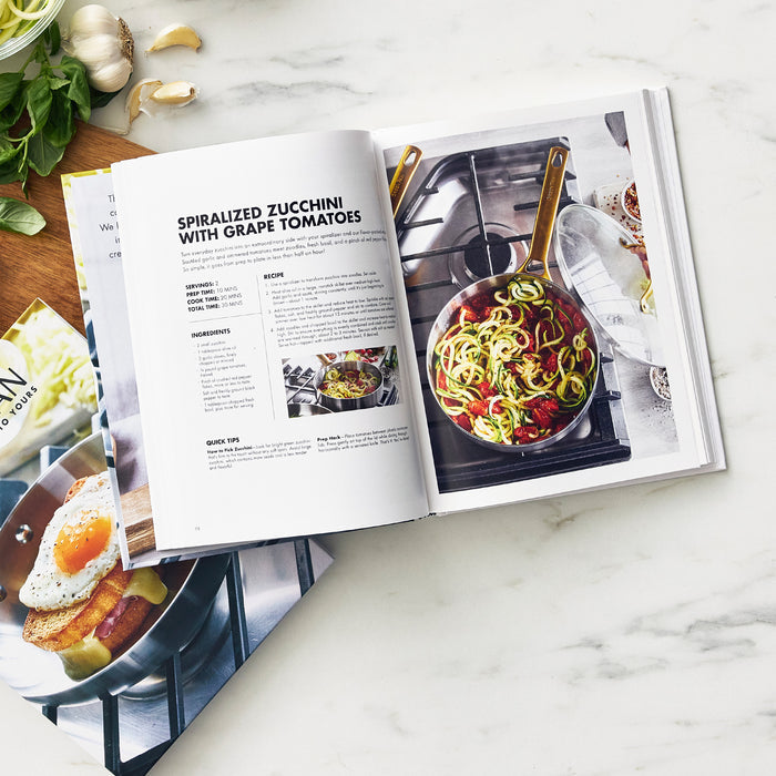 An open Cook's Journal Cookbook, Vol. 2 rests on a marble surface, displaying a spiralized zucchini with grape tomatoes recipe and a photo of zucchini noodles and tomatoes cooking in a skillet, with garlic, basil, and other cookbooks nearby.