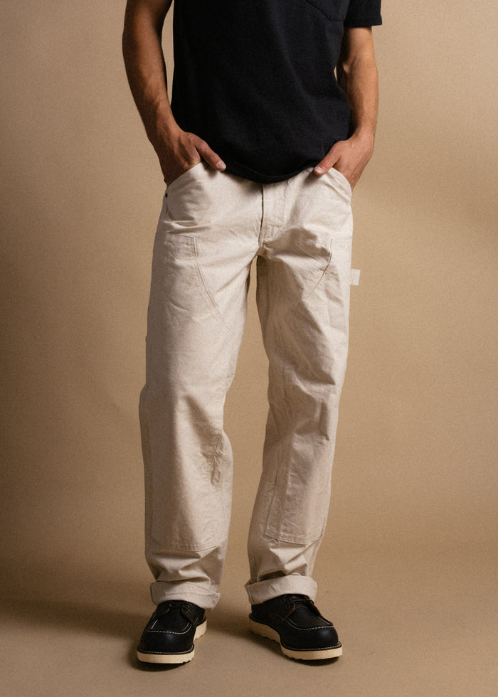 Man in black shirt and beige pants stands with hands in pockets, wearing black shoes, against a plain tan background.