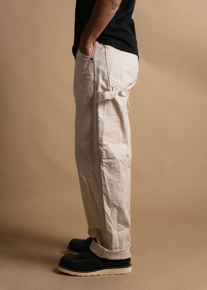 Man in beige cargo pants and black shoes stands sideways against a tan background, hands in pockets, wearing a black shirt.