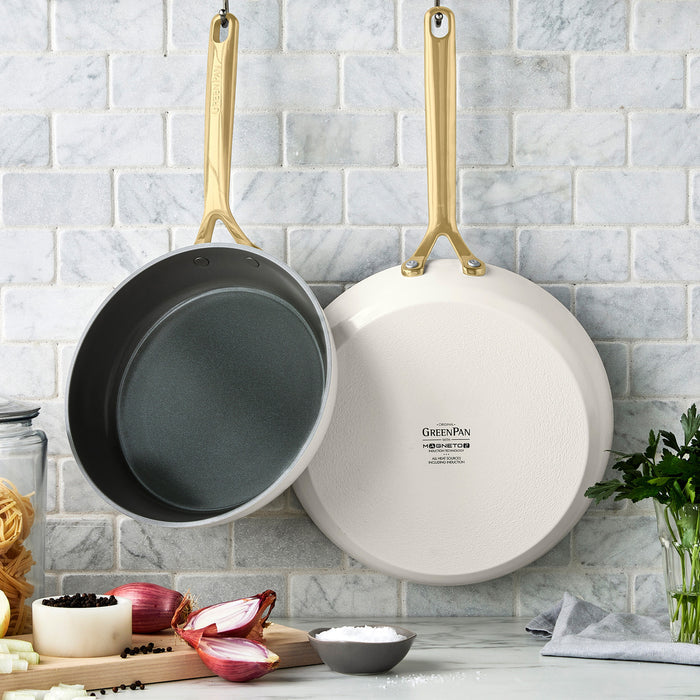 Two ceramic nonstick frypans with champagne handles hang on a tiled wall above fresh herbs.
