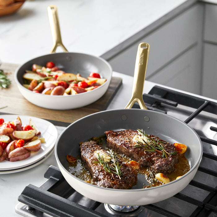 Two steaks cook in a green ceramic nonstick frypan, with potatoes in a cream-colored pan nearby on the stove.