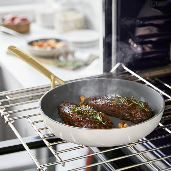 Two cooked steaks with herbs in a cream-colored ceramic nonstick frypan set with champagne handles.