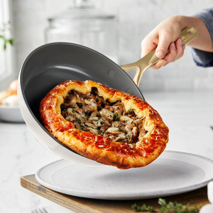 Hand sliding mushroom galette from ceramic nonstick frypan onto a plate in a bright kitchen.