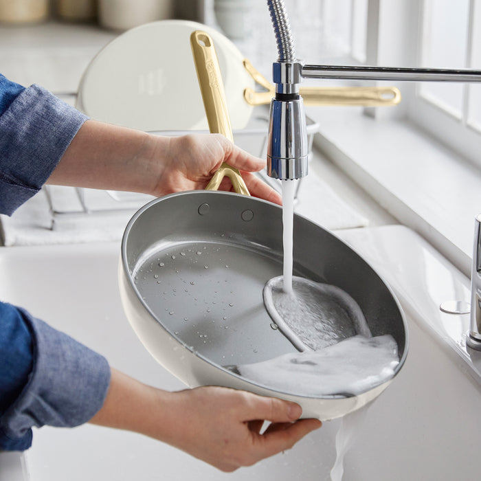 Ceramic frying pan set with champagne handles in cloud cream being rinsed under a kitchen faucet.