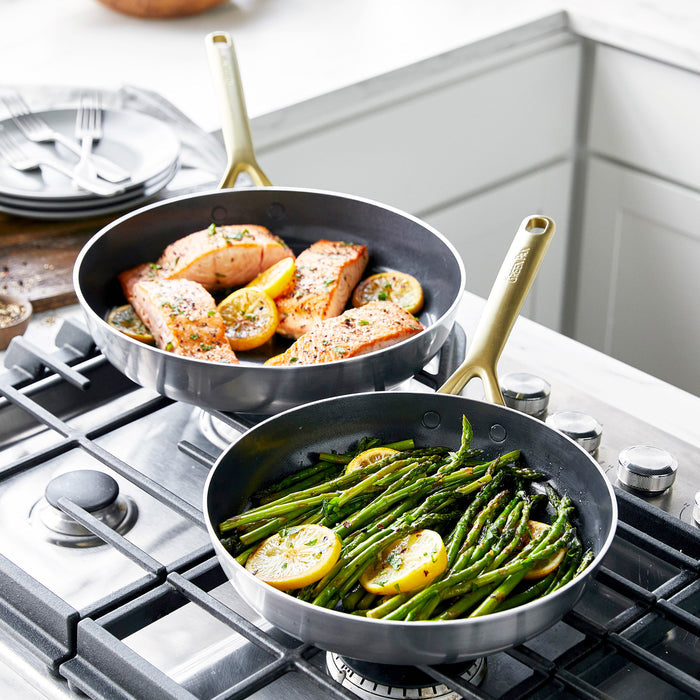 Stainless steel frypan set with champagne handles, two pans cooking salmon and asparagus on a kitchen stovetop.