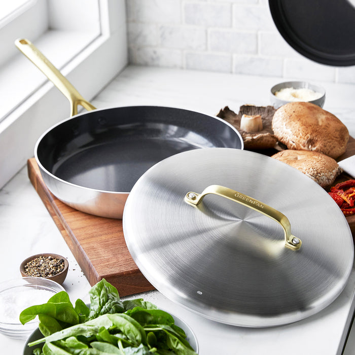Stainless steel frypan with lid and champagne handles on a kitchen counter, next to fresh spinach and mushrooms.