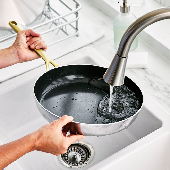 Stainless steel frypan with champagne handles and lid being rinsed under a kitchen faucet by a white sink.