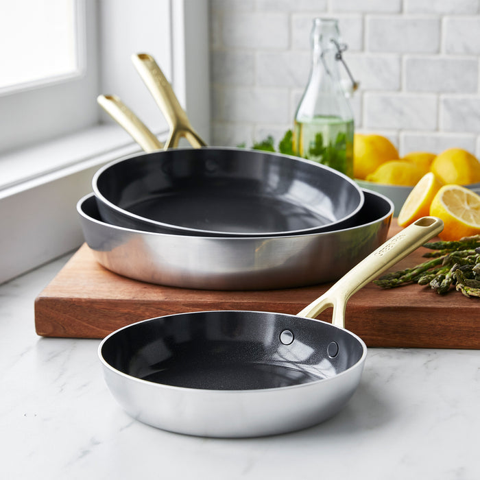 Three stainless steel frypans with champagne handles, stacked on a wooden board with lemons and herbs in the background.