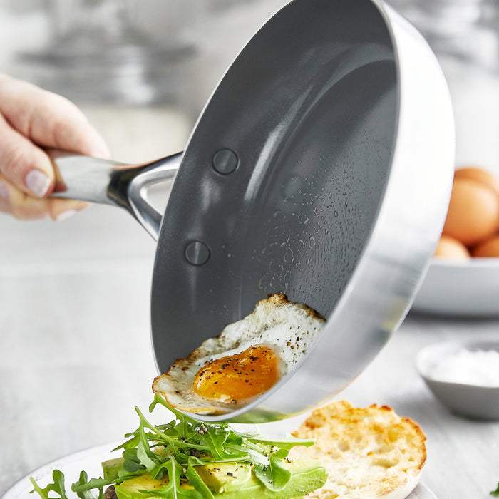 Hand tilting stainless steel frypan to slide a fried egg onto avocado arugula toast.