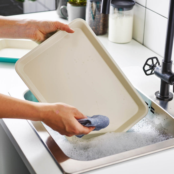 Hand washing a green ceramic nonstick cookie sheet set, two pieces, with a sponge in a soapy sink.