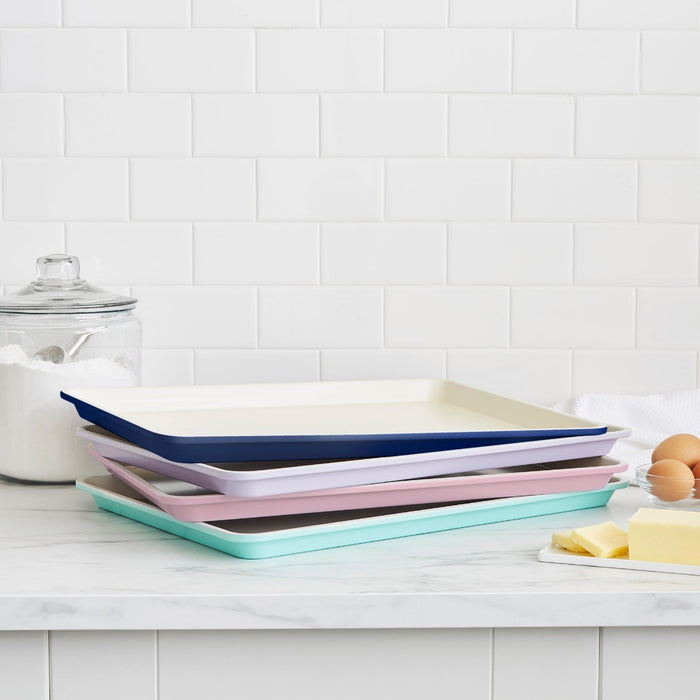 Set of four green ceramic 13x9 inch cookie sheets stacked on a kitchen counter with baking ingredients behind.
