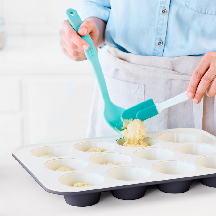 Scooping muffin batter into a black GreenLife ceramic nonstick muffin pan with a spatula in a bright kitchen.