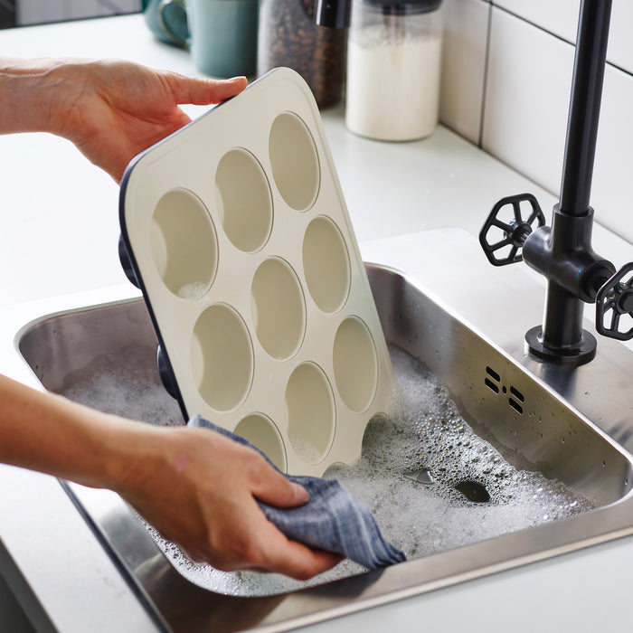 Washing a black GreenLife ceramic muffin pan in a soapy sink beside a black faucet.