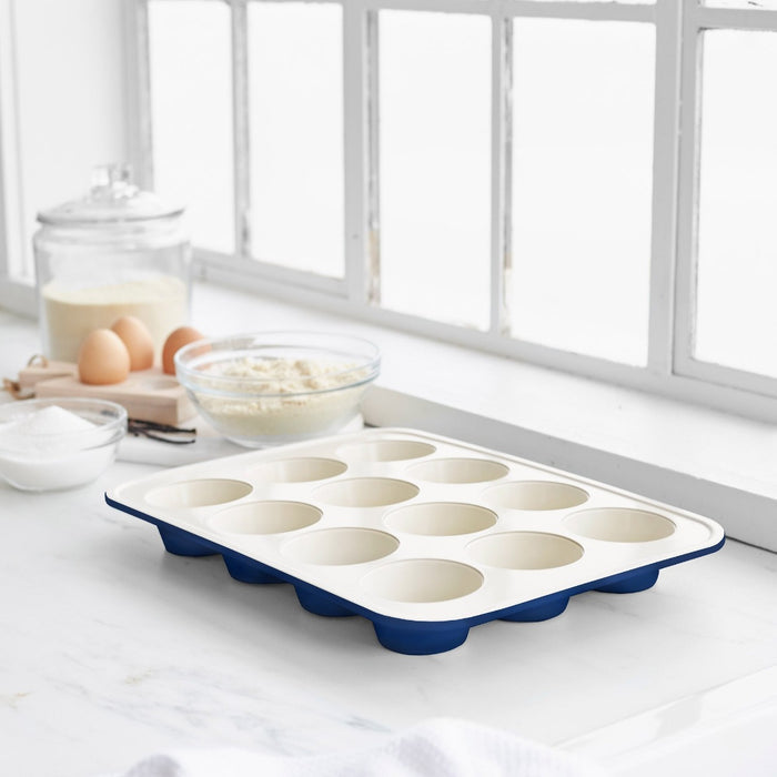 Navy ceramic nonstick muffin pan on a kitchen counter with baking ingredients, sunlight streaming through a window.