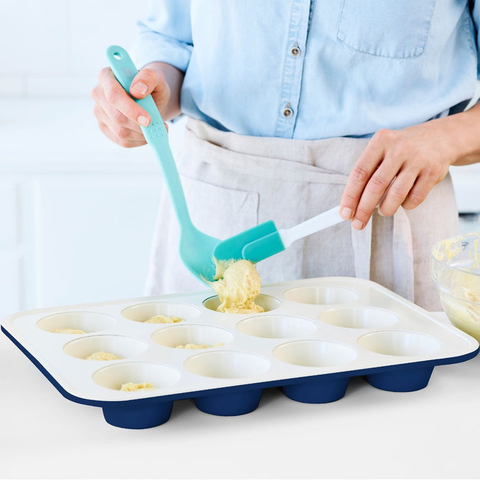 Person using a blue spatula to fill a navy GreenLife ceramic muffin pan with batter in a well-lit kitchen.