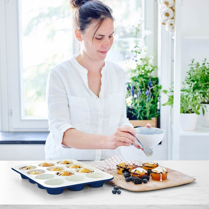Woman icing muffins beside a navy GreenLife ceramic muffin pan on a kitchen counter with houseplants.