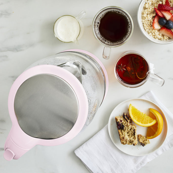 Pink glass electric kettle with tea cups, milk, oatmeal, fruit, and bread on a white tabletop.