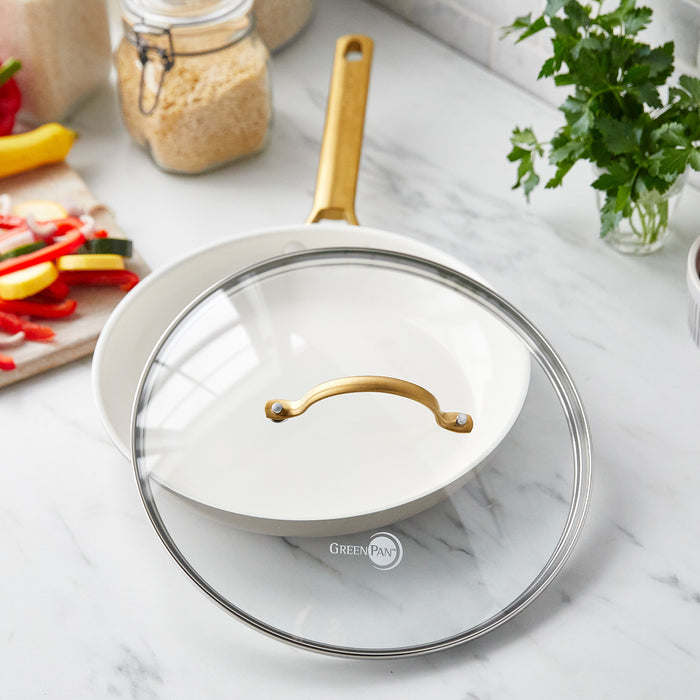 Glass lid on white skillet with matte gold handle, surrounded by assorted vegetables and herbs on a marble surface.