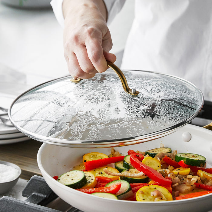 Glass pan lid with matte gold handle being lifted from sautéed vegetables in a pan.