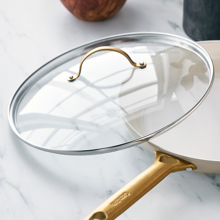 Glass lid with matte gold handle resting on a white pan, set on a marble countertop.