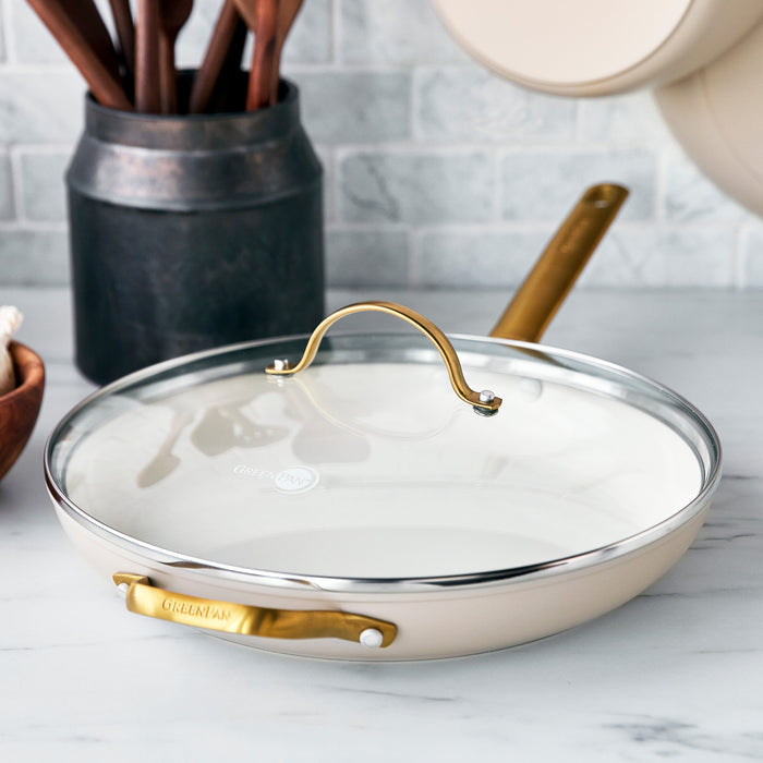 Green frying pan with matte gold handle and glass lid on a white countertop.