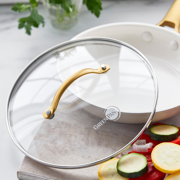 Glass saucepan lid with stainless rim on a cutting board beside a white pan holding sliced vegetables and fresh herbs.