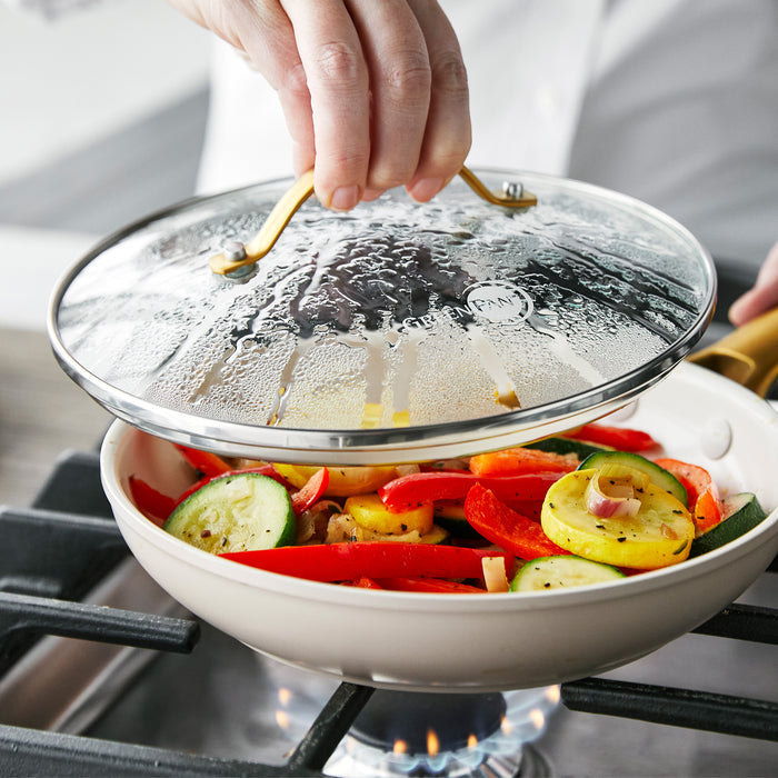 A hand lifts a glass GreenPan lid from a pan with sautéed vegetables on a stovetop.