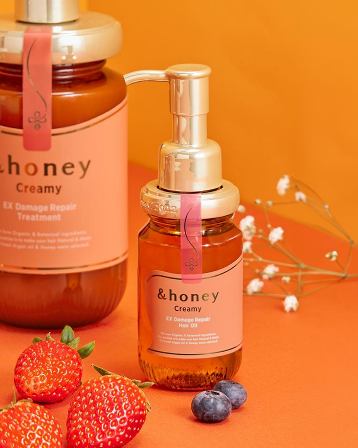 Two bottles of &honey Creamy EX Damage Repair hair oil and treatment, displayed on an orange background with fresh strawberries, blueberries, and delicate white flowers, representing authentic K-beauty skincare available at Sukoshi Mart.