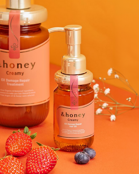 Two bottles of &honey Creamy EX Damage Repair hair oil and treatment, displayed on an orange background with fresh strawberries, blueberries, and delicate white flowers, representing authentic K-beauty skincare available at Sukoshi Mart.