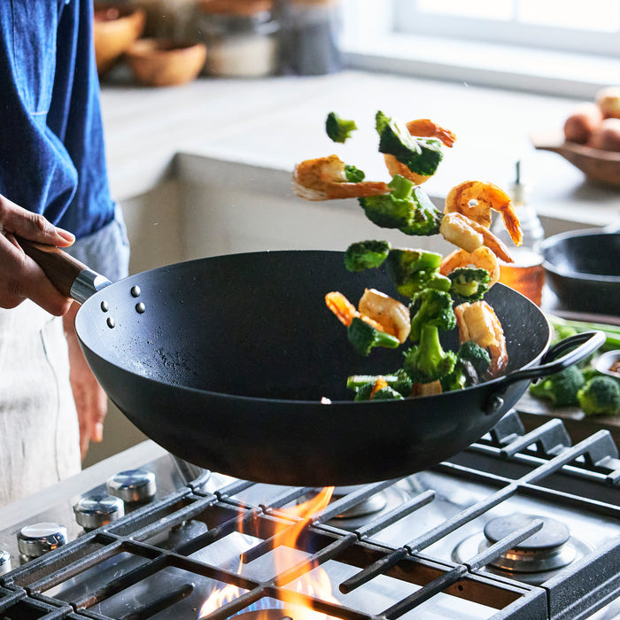 Person cooking shrimp and broccoli in a wok on a gas stove with flames.