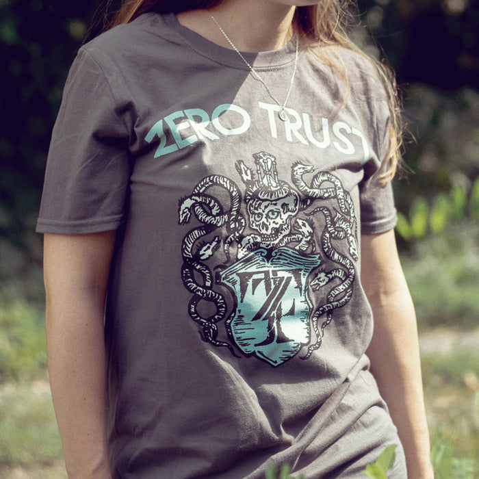 Charcoal colored short sleeve shirtwith ZERO TRUST written across the top. Below the text is a drawing of a skeleton with snakes surrounding it, drawn in white. Below the skeleton is a crest that has the letters Z and T written inside it. An individual is modeling the shirt and standing outside on grass