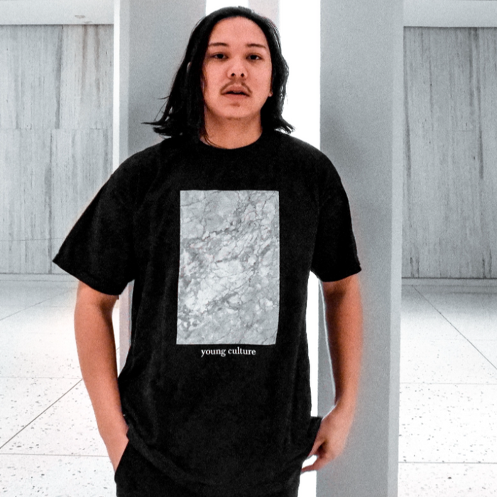 Black short sleeve shirt with a rectangle design filled with white and grey marble pattern. Below that there is small white text that says young culture. An individual is modeling the shirt and standing inside against a white pole.
