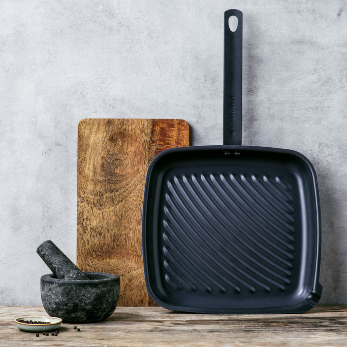 Square carbon steel grill pan with long handle on wooden surface, shown with accompanying accessories.