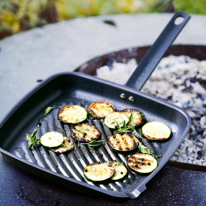 Zucchini slices cooking in a Merten  Storck carbon steel square grill pan over an outdoor flame.