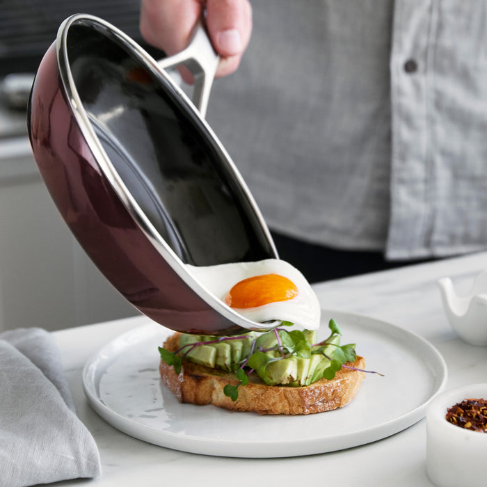 Sunny-side-up egg sliding from a Merten  Storck 9.5 steel core frypan onto avocado toast with leafy greens.