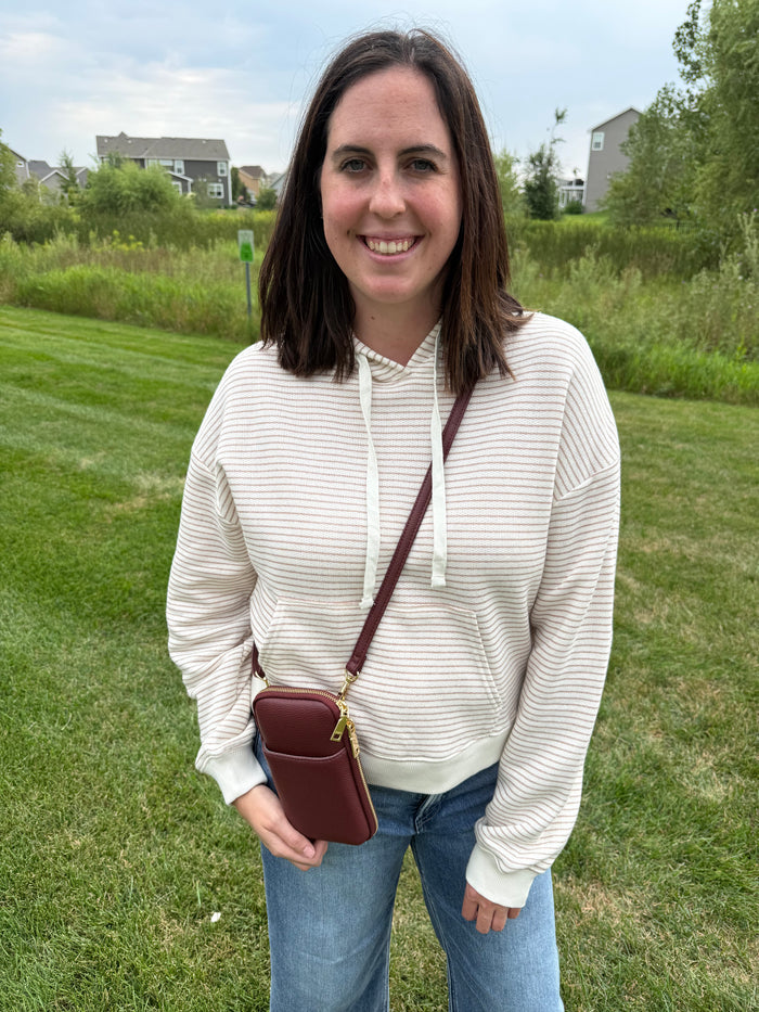 A woman wearing a cream and taupe horizontally striped hoodie, blue jeans, and carrying a small plum crossbody phone pouch with gold hardware.