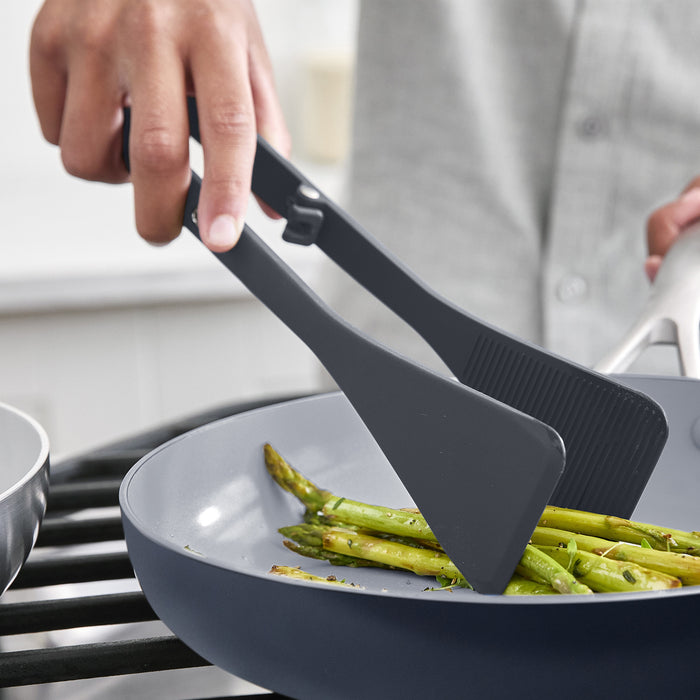 Black silicone tongs picking up asparagus from a pan, part of the gray 4-piece utensil and tool rest set.