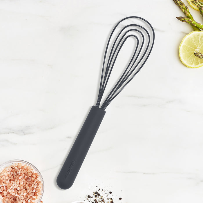 Gray silicone flat whisk with black handle on marble surface, next to salt, pepper, lime wedges, and asparagus.