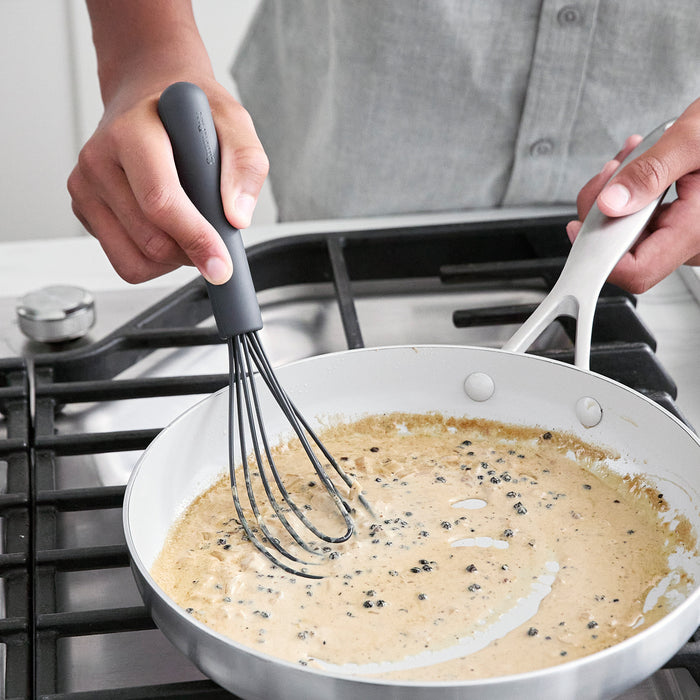 Gray silicone flat whisk stirring creamy peppercorn sauce in a pan on a stovetop.
