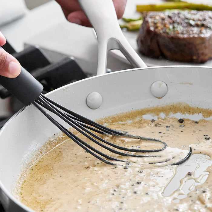 Gray platinum silicone flat whisk blending sauce in a white pan, with steak and asparagus in the background.