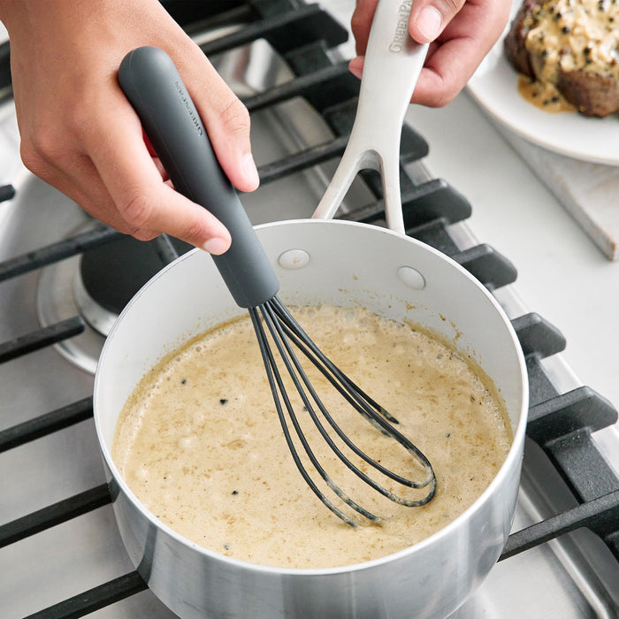 Gray platinum silicone flat whisk stirring creamy sauce in a saucepan, with a plate of steak nearby.